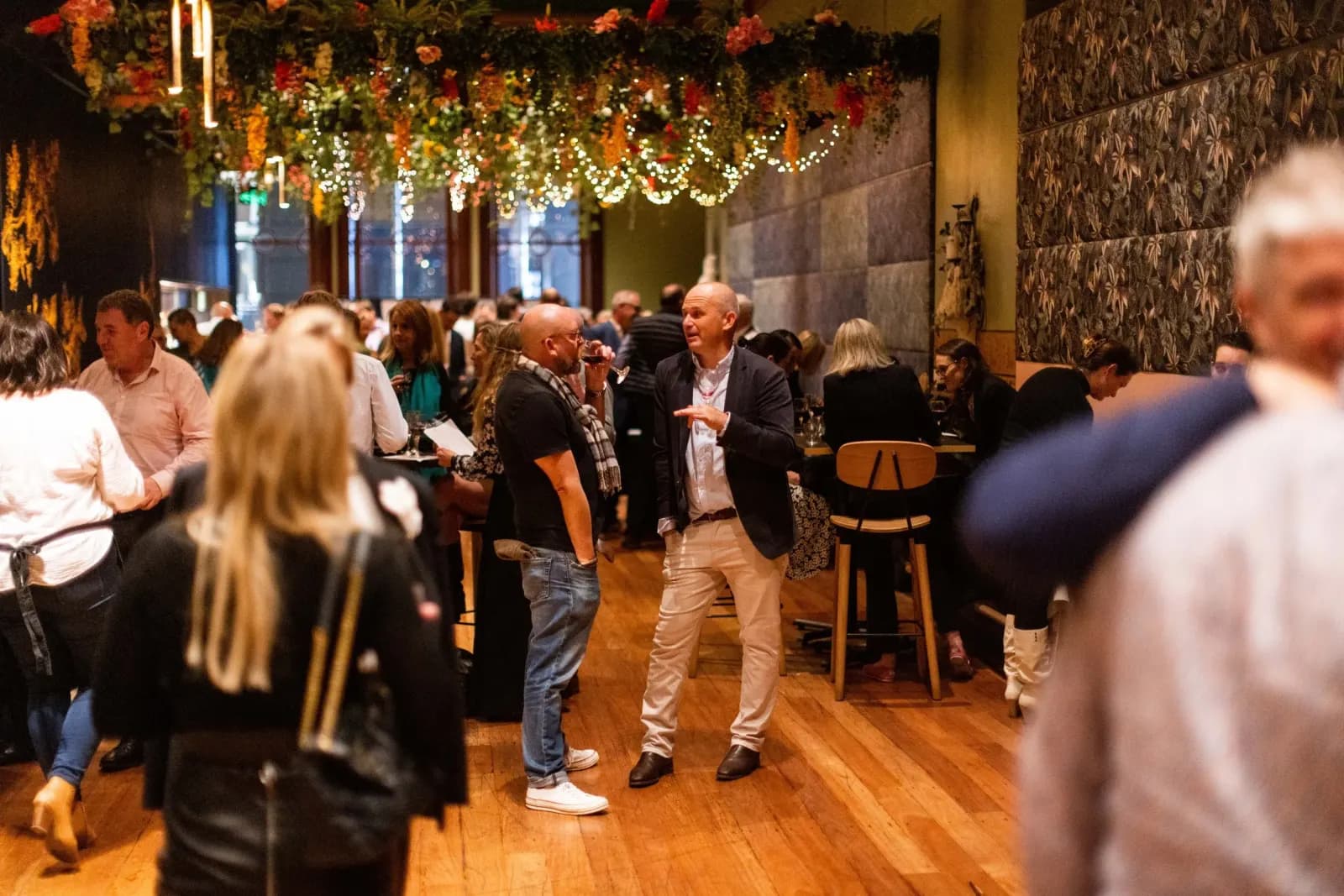 A full room at Gather & Yarn, guests talking under hanging greenery and lights